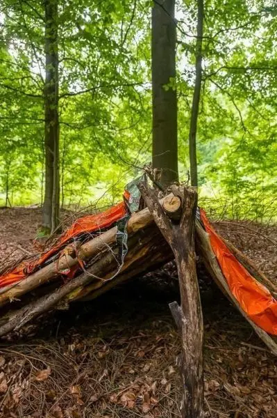 Tarp-Notunterkunft aus Ästen und einer orangefarbenen Plane unter Bäumen